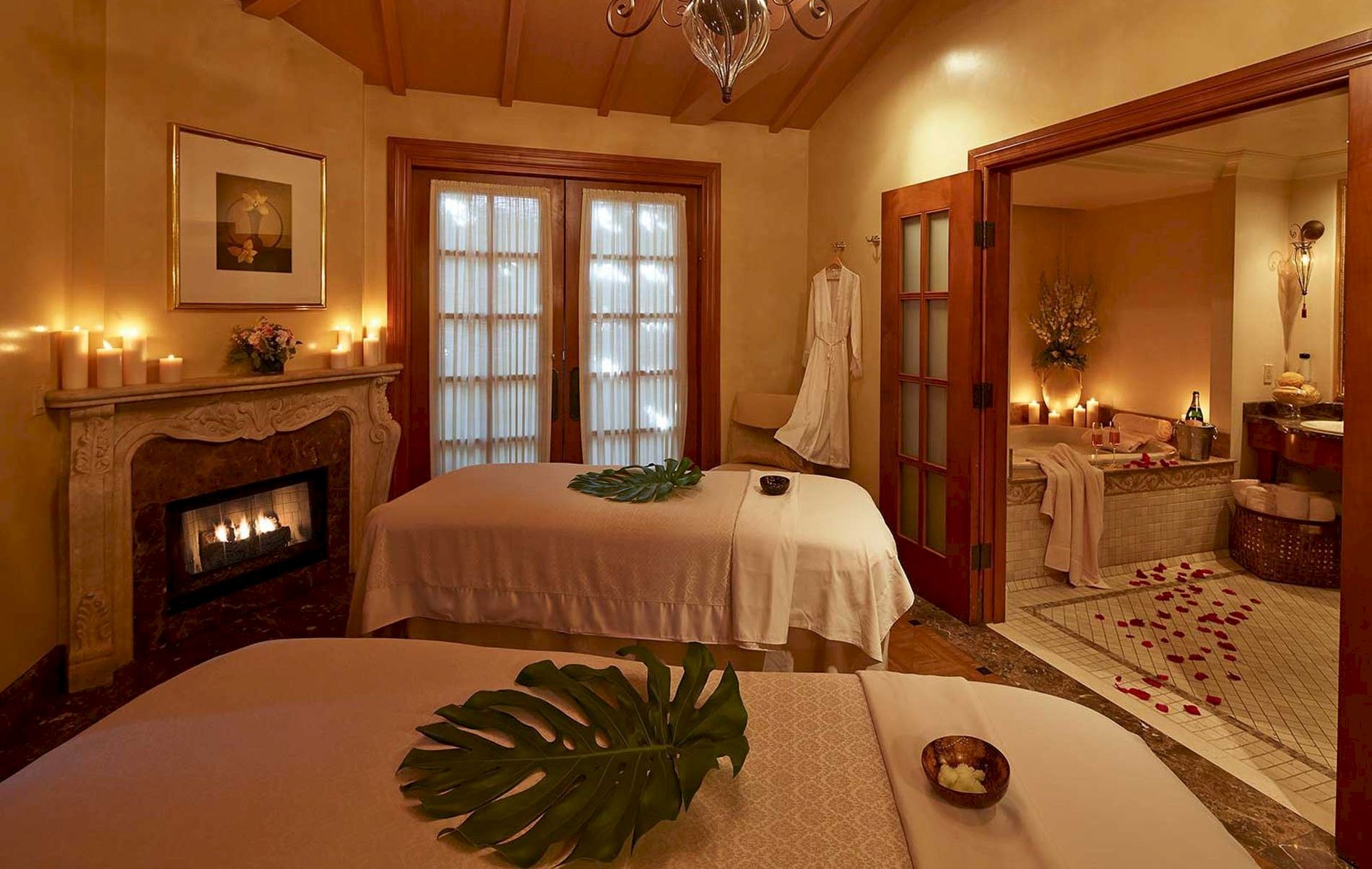 A warm, candlelit spa room with two beds, a cozy fireplace, soft lighting, tropical leaf decor, and an adjoining mirror-lit bathroom area.
