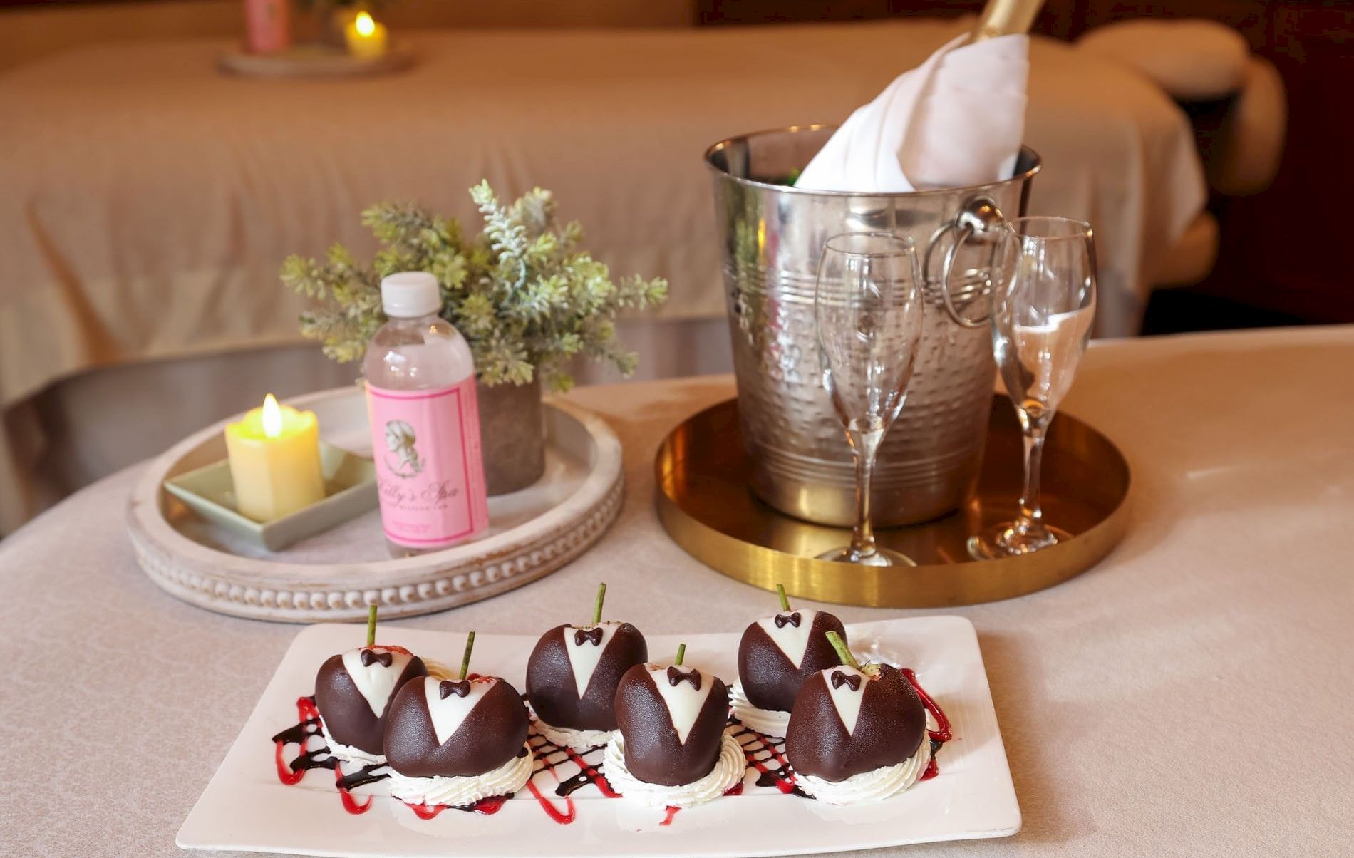 Assorted items on a spa-like setup: a tray with candles and flowers, a bottle of pink lotion, ice bucket with a bottle and glasses, and chocolate desserts on a plate.
