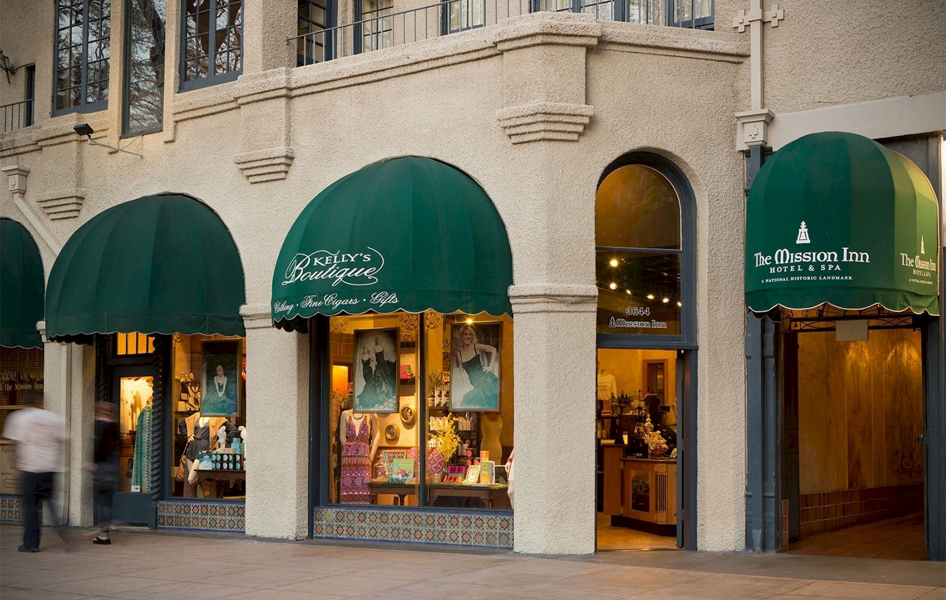 A shopfront with three green awnings on a beige building; people walking by and store windows displaying fashion items.