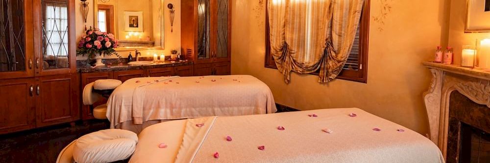 A warm, cozy spa room with two massage beds, soft lighting, fragrant candles, floral decor, and ornate wooden furniture.