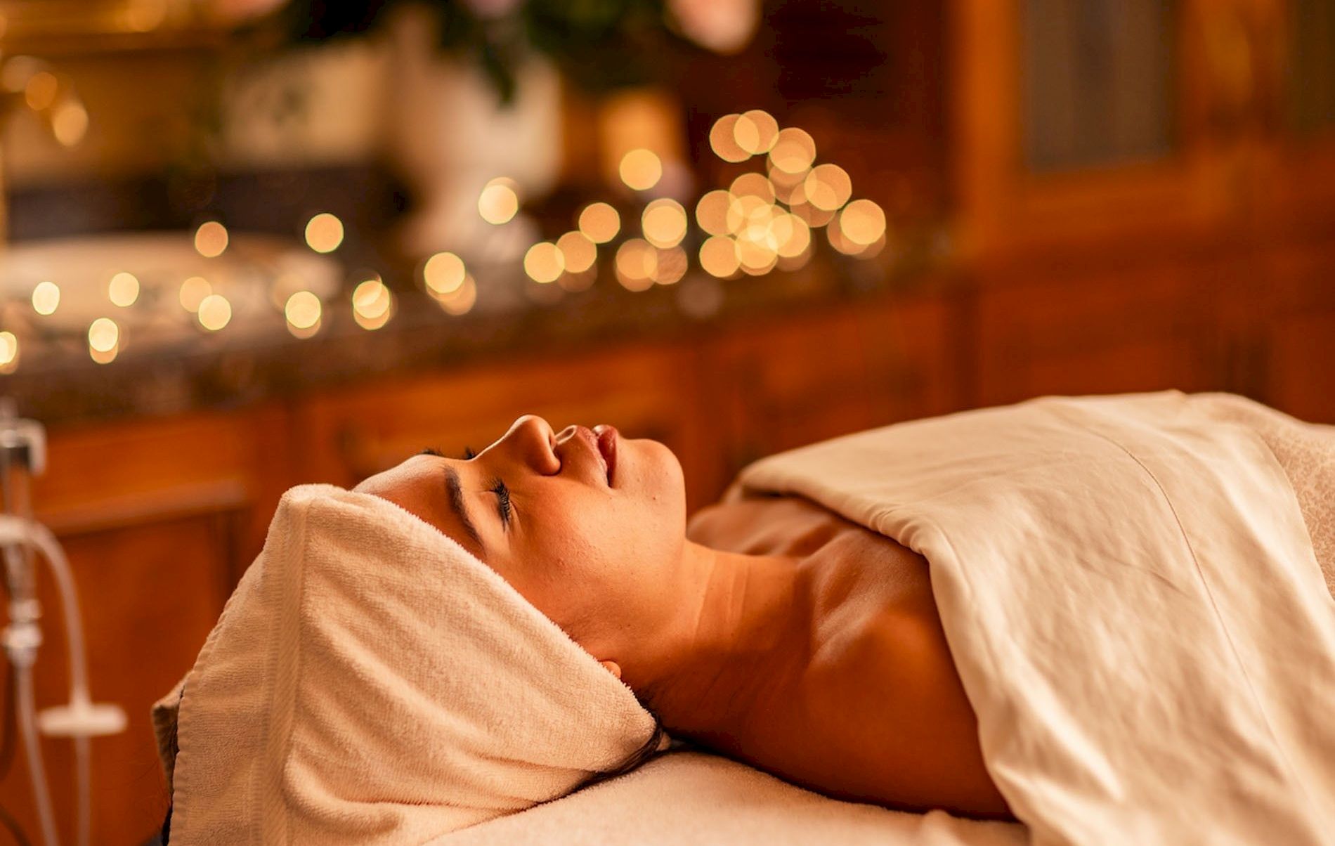 A person lying on a treatment bed with a towel wrapped around their head, relaxing in a softly lit spa setting with warm, out-of-focus lights.
