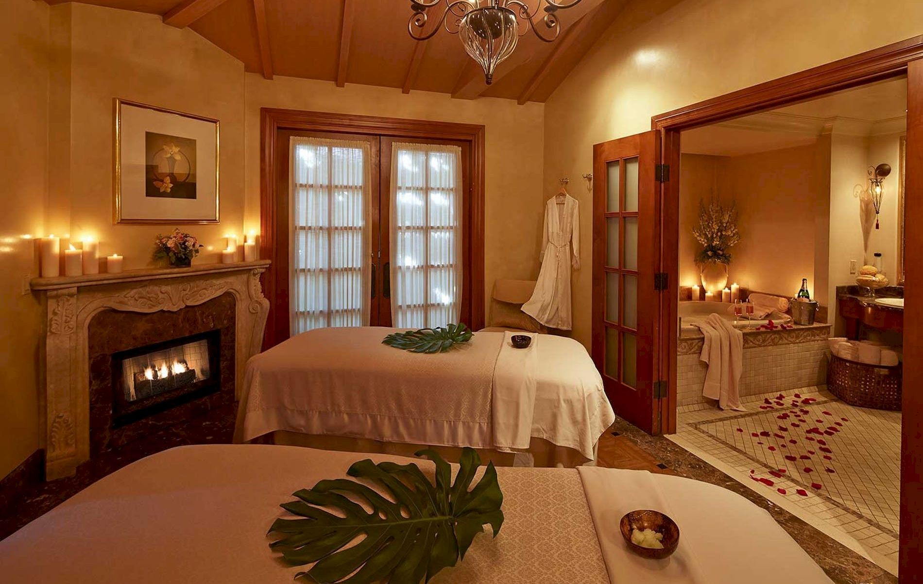A cozy, warmly lit spa-like room with two massage beds, candles by the fireplace, and tropical leaf decor, creating a tranquil retreat atmosphere.