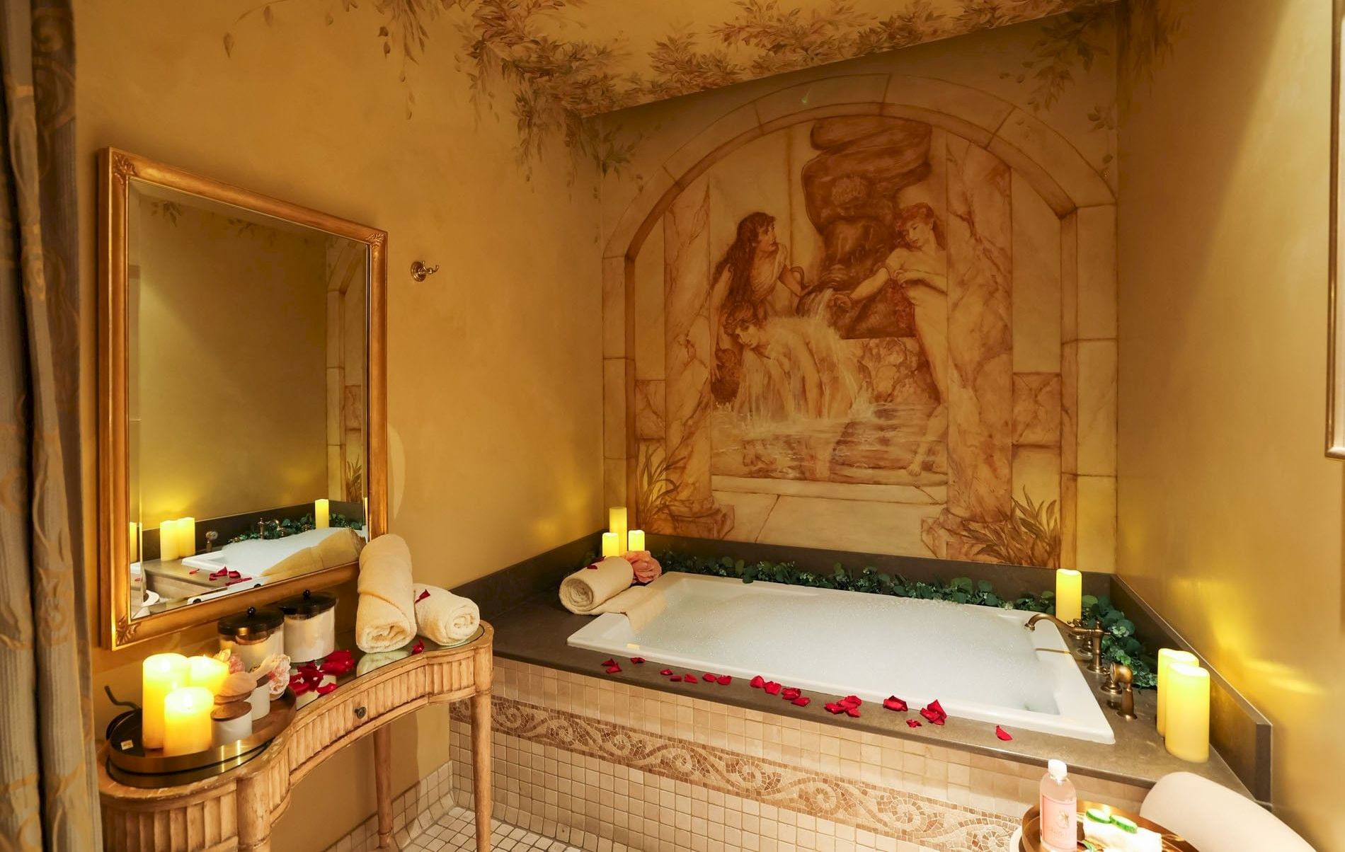 A cozy, candlelit bathroom with a sunken tub, mosaic wall art, and rose petals scattered around.