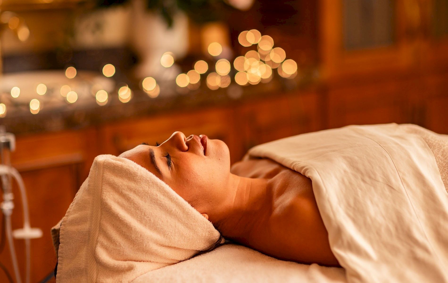 A person is lying on a bed with a towel wrapped around their head, in a cozy dimly lit room with soft bokeh lights in the background.