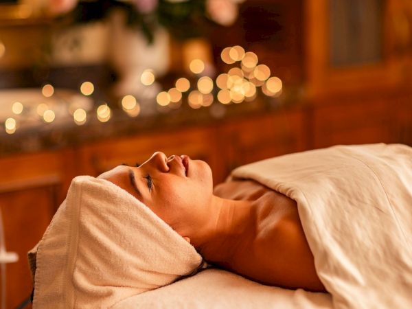 A woman in a spa or clinic lies on a bed with a towel wrapped around her head, eyes closed, soft warm lights glowing in the background.