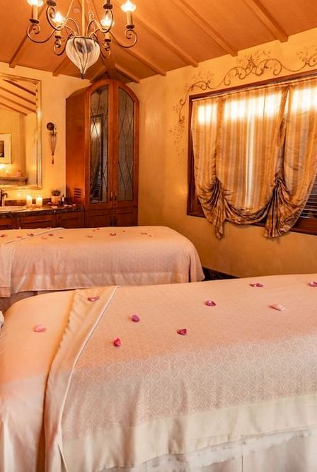 A cozy, warmly lit spa room with two massage tables, soft cream linens, rose petals, wood furniture, and a decorative window curtain.