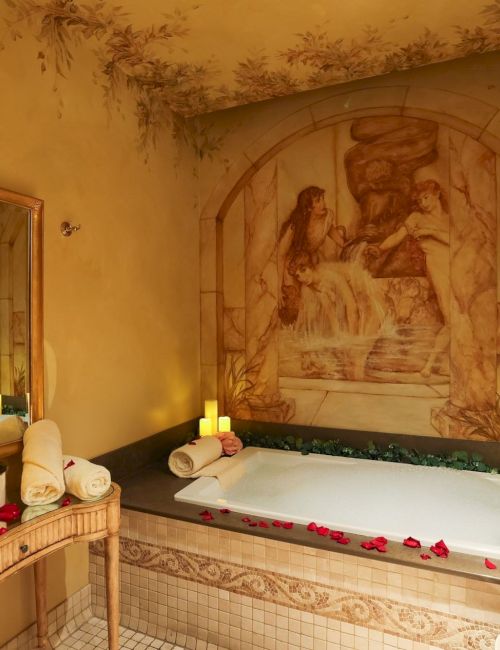 A cozy, candlelit bathroom with a large spa tub, rose petals, soft lighting, a wall mural, and a tiled dressing area with a mirror and towels.