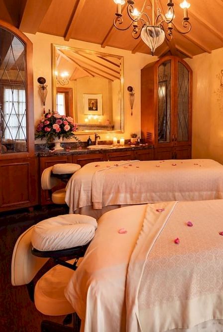 A warm, vintage spa-like room with two massage beds, soft lighting, wooden cabinets, ornate drapes, candles, and a floral decor ambiance.