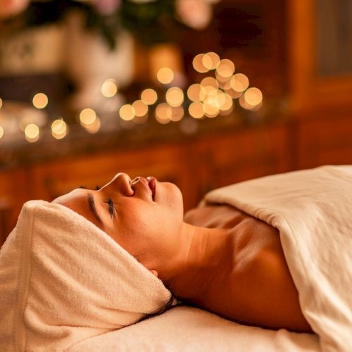 A woman lies on a spa table with a towel wrapped around her head, eyes closed, in a softly lit, cozy setting with warm bokeh lights in the background.