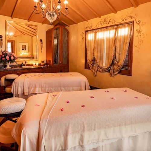 A cozy spa room with two massage beds, warm lighting, wooden furniture, and soft rose petals; a chandelier, curtains, and a fireplace add a romantic, luxurious vibe.