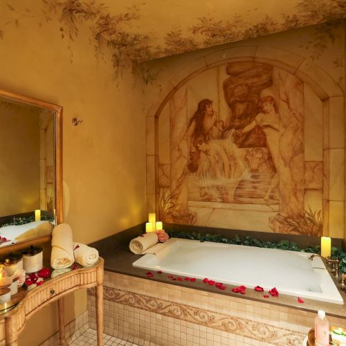 A candle-lit bathroom with a large tub, rose petals, wall mural, and a mirror reflecting a cozy, warm spa vibe.