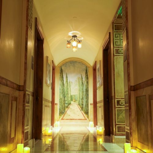 A long, elegant hallway lined with glowing candles, marble floors, and ornate walls leading to a serene garden mural at the end.