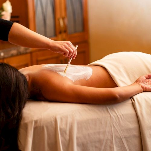 A massage therapist applies white lotion to a woman's back with a stick-like tool, as she lies face down on a massage table under warm lighting.