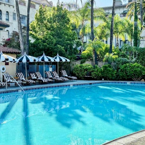 A sunny hotel pool surrounded by palm trees, lounge chairs, and striped umbrellas. It&rsquo;s a relaxing resort scene.