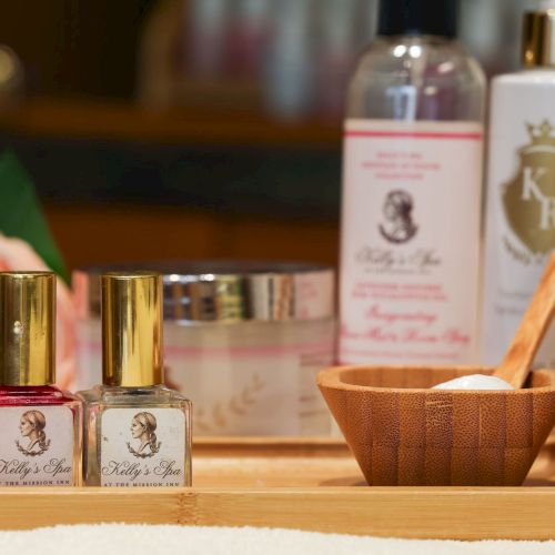 A vanity scene with pink nail polish bottles front and center, small jars, a wooden bowl with a spoon, and blurred skincare bottles in the background.