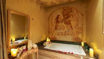 A cozy spa bathroom with a candlelit tub, soft lighting, and rose petals arranged around the tub, plus a mural backdrop.