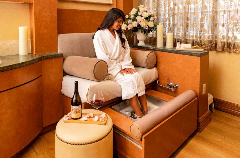 A woman in a white robe sits on a spa chair, feet soaking, with a bottle of wine, glasses, and candles nearby in a cozy treatment room. ending sentence.