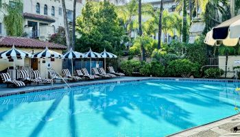 A sunny hotel pool scene with blue water, lounge chairs, striped umbrellas, palm trees, and surrounding white buildings.