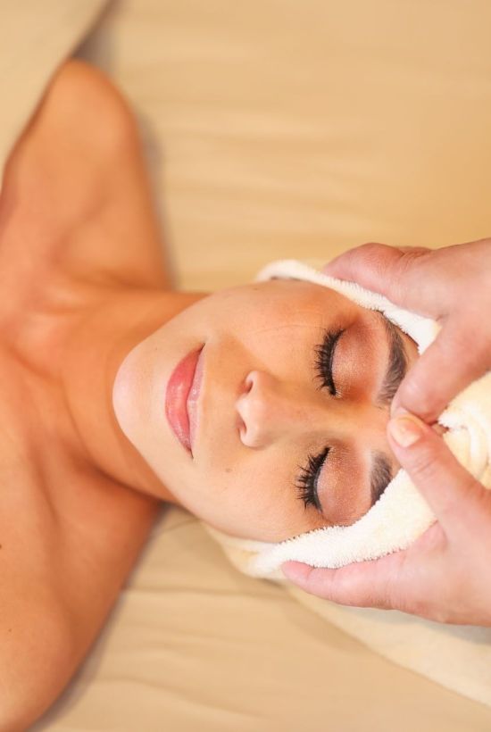A woman at a spa receiving a facial, eyes closed, towel wrapped around her head, lying on a bed.