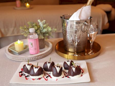 A table with chocolate-dipped strawberries on a white plate, a candle, a pink-labeled bottle, flowers, and a champagne bucket with glasses in a cozy setting.