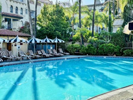 A vibrant hotel pool area with clear blue water, striped lounge chairs under umbrellas, palm trees, and lush greenery surrounding the pool.