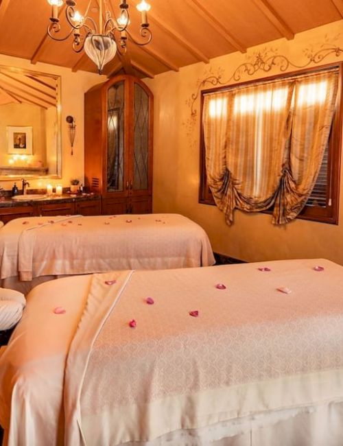 A cozy spa-like room with two massage beds, warm amber lighting, ornate wood furniture, floral decor, a chandelier, and a window with draped curtains.